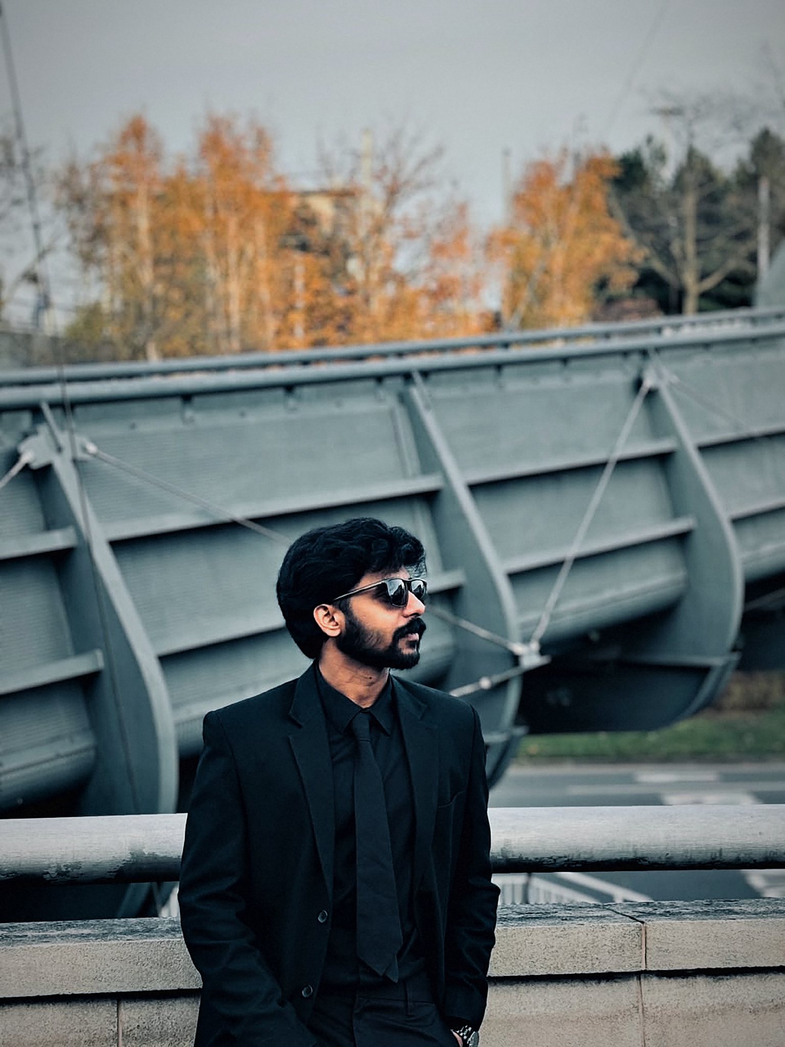 Arun in a black suit and sunglasses, looking out over an autumn bridge