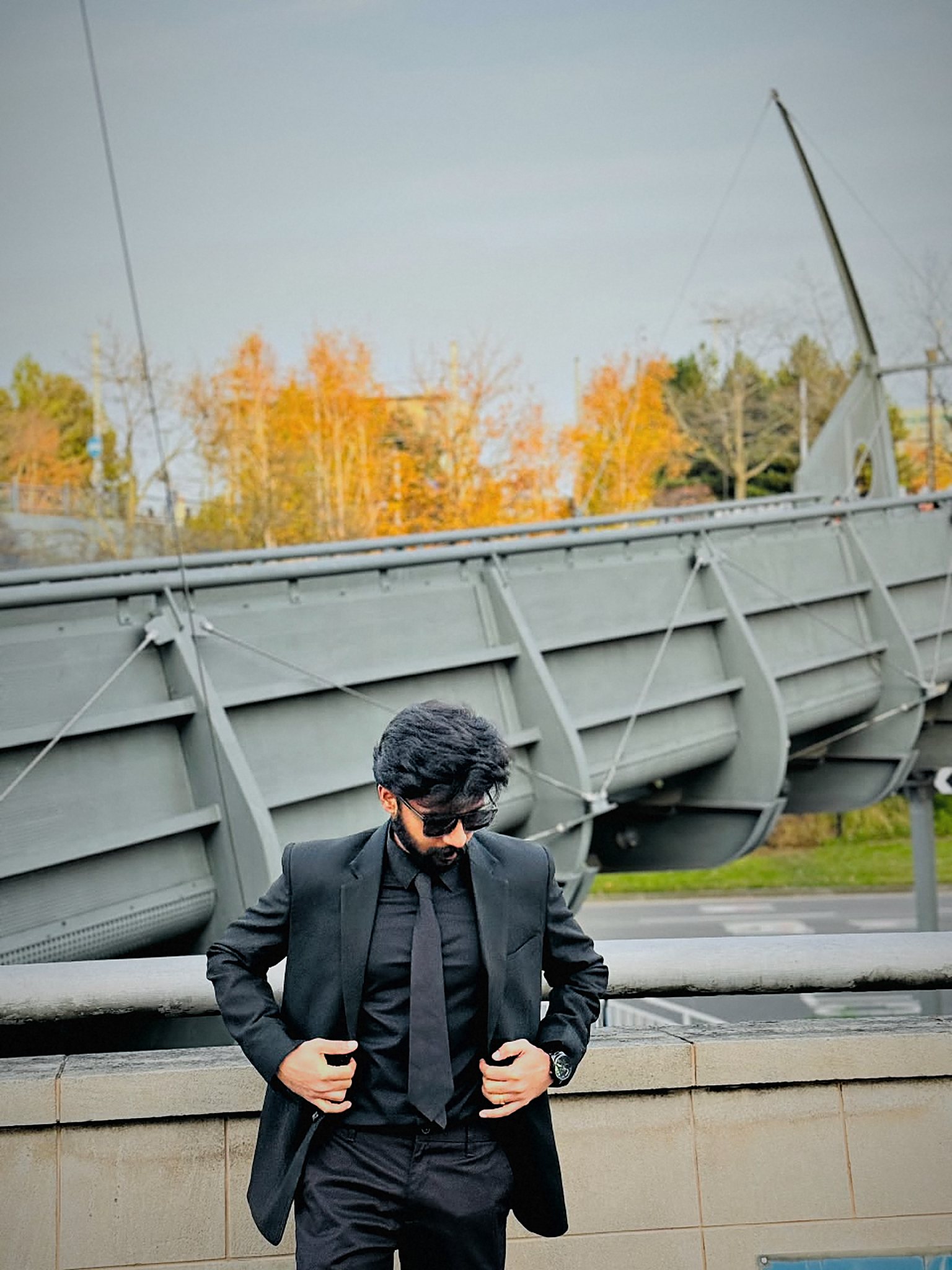 Arun adjusting his suit jacket, eyes down, under a steel footbridge