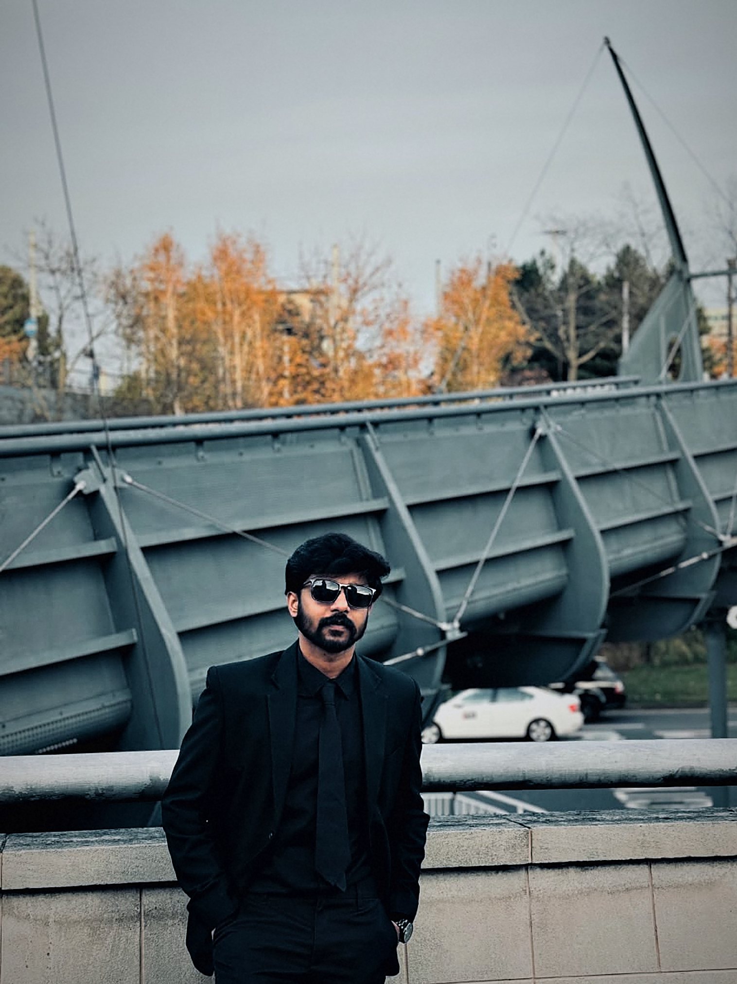Arun leaning on a stone wall, hands in pockets, in sunglasses