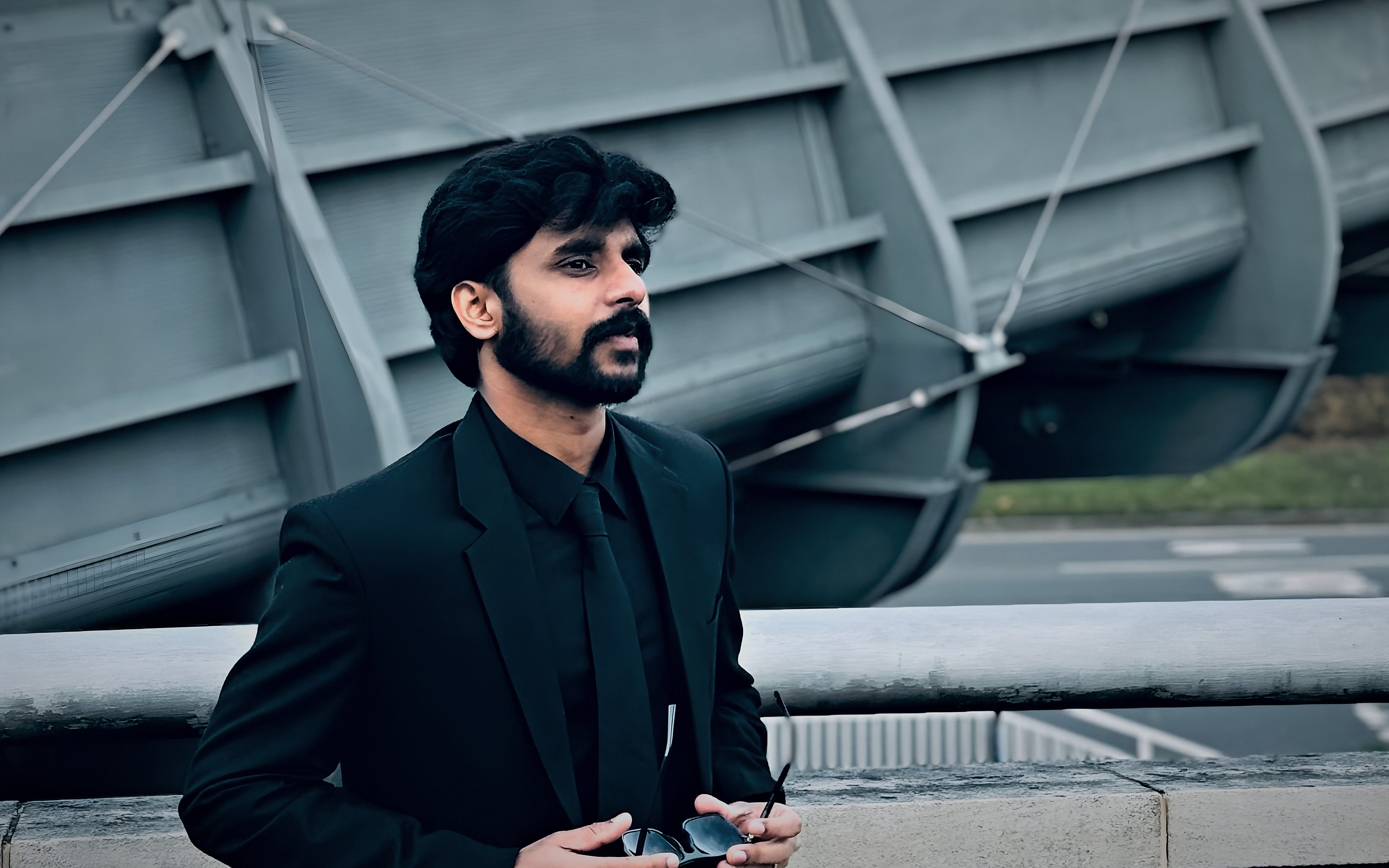 Arun Reena Raju in a black suit, standing in front of a steel footbridge